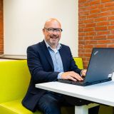 Een lachende man werkt op een laptop aan een tafel met een gele bank.